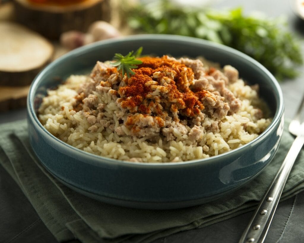 A blue bowl filled with cooked rice and seasoned ground meat, offering 30g protein and only 250 calories, topped with red paprika and parsley, sits on a green cloth with a blurred background of herbs.