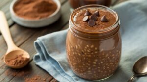 A glass jar filled with Chocolate Brownie Batter Overnight Oats, topped with cocoa powder and chocolate chips, sits on a blue cloth with a wooden spoon and a bowl of cocoa powder nearby on a rustic wooden table.
