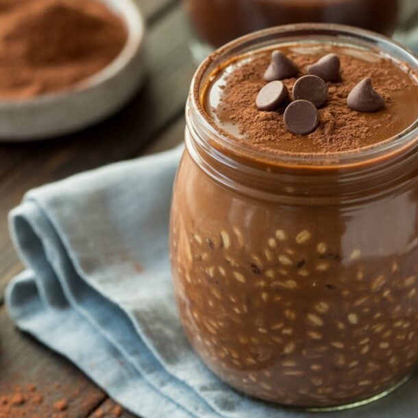 A glass jar filled with Chocolate Brownie Batter Overnight Oats, topped with cocoa powder and chocolate chips, sits on a blue cloth with a wooden spoon and a bowl of cocoa powder nearby on a rustic wooden table.