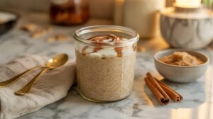 A glass jar filled with Cinnamon Roll Overnight Oats, topped with cinnamon, sits on a marble surface next to two gold spoons, a napkin, a bowl of brown sugar, and two cinnamon sticks. Warm, soft lighting creates a cozy atmosphere.