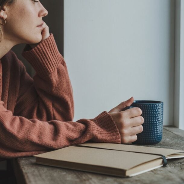 A person in a pink sweater sits by a window, resting their chin on their hand and holding a blue mug. An open notebook for tracking signs of cortisol imbalance lies on the wooden table beside them, with a small plant in the corner.