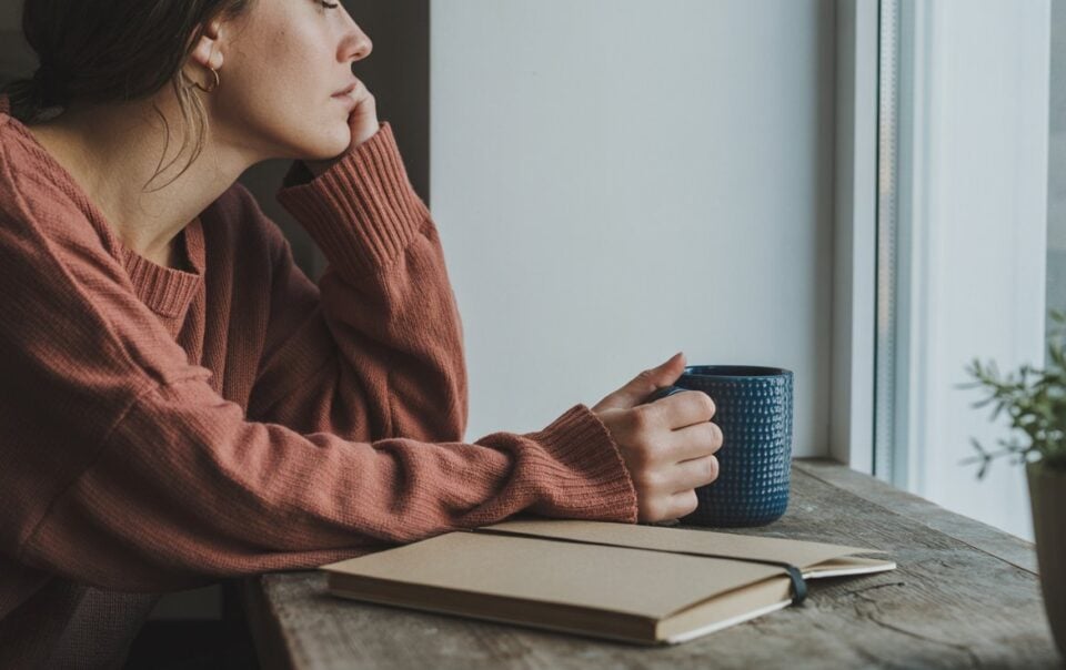 A person in a pink sweater sits by a window, resting their chin on their hand and holding a blue mug. An open notebook for tracking signs of cortisol imbalance lies on the wooden table beside them, with a small plant in the corner.