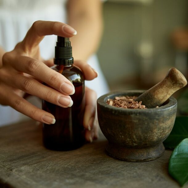 A person’s hand holds a brown glass spray bottle of DIY Magnesium Spray Deodorant beside a stone mortar filled with pink salt and a pestle, on a rustic wooden table with a lemon and green leaves nearby.