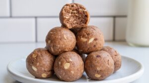 A pyramid stack of seven date protein balls sits on a white plate. The top ball has a bite taken out, revealing a soft, nutty interior. The background is a white tiled wall and a blurred glass of milk.