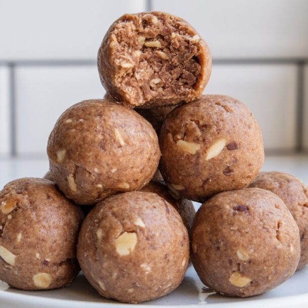 A pyramid stack of seven date protein balls sits on a white plate. The top ball has a bite taken out, revealing a soft, nutty interior. The background is a white tiled wall and a blurred glass of milk.