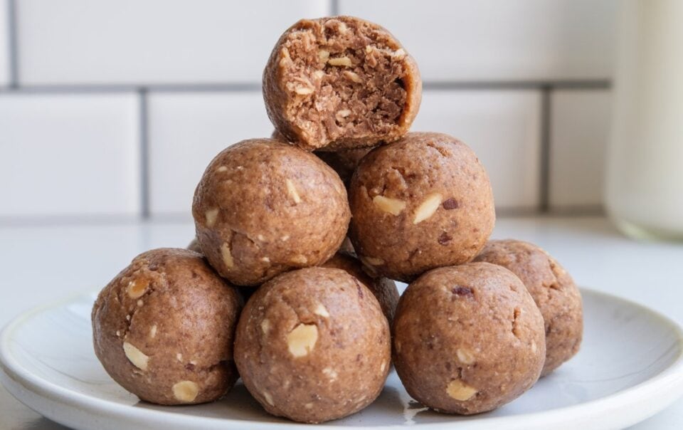 A pyramid stack of seven date protein balls sits on a white plate. The top ball has a bite taken out, revealing a soft, nutty interior. The background is a white tiled wall and a blurred glass of milk.