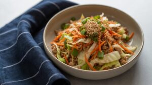 A bowl of egg roll in a bowl—stir-fried ground meat, shredded cabbage, carrots, and green onions topped with sesame seeds—rests beside a dark blue cloth napkin with white stripes on a light surface.