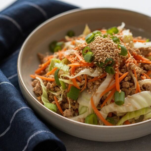 A bowl of egg roll in a bowl—stir-fried ground meat, shredded cabbage, carrots, and green onions topped with sesame seeds—rests beside a dark blue cloth napkin with white stripes on a light surface.