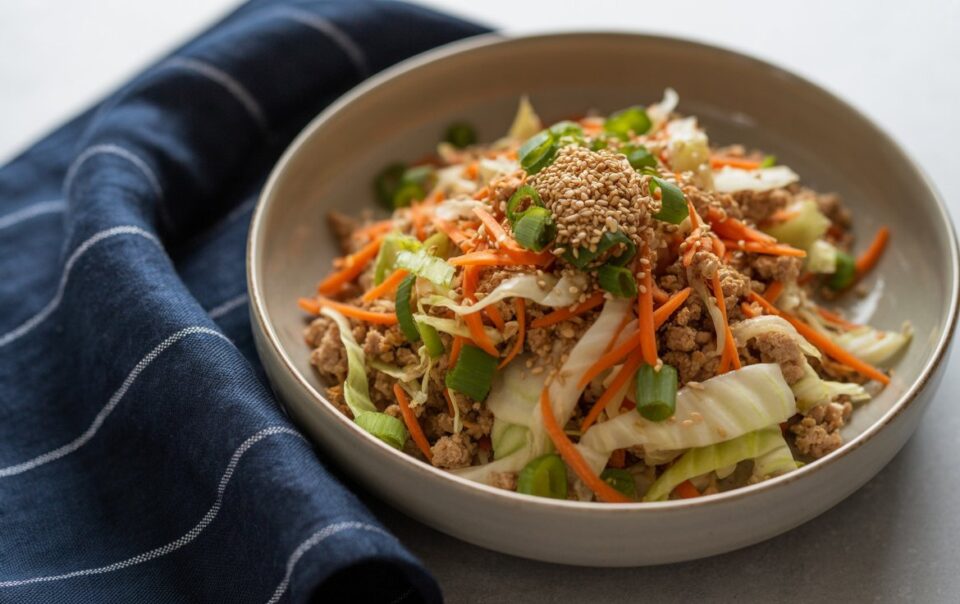 A bowl of egg roll in a bowl—stir-fried ground meat, shredded cabbage, carrots, and green onions topped with sesame seeds—rests beside a dark blue cloth napkin with white stripes on a light surface.