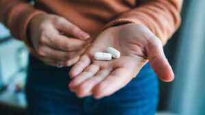 A person holds two large white Foundayo weight loss pills in the palm of their hand, with their other hand reaching toward the pills. The individual is wearing a brown sweater and blue jeans.