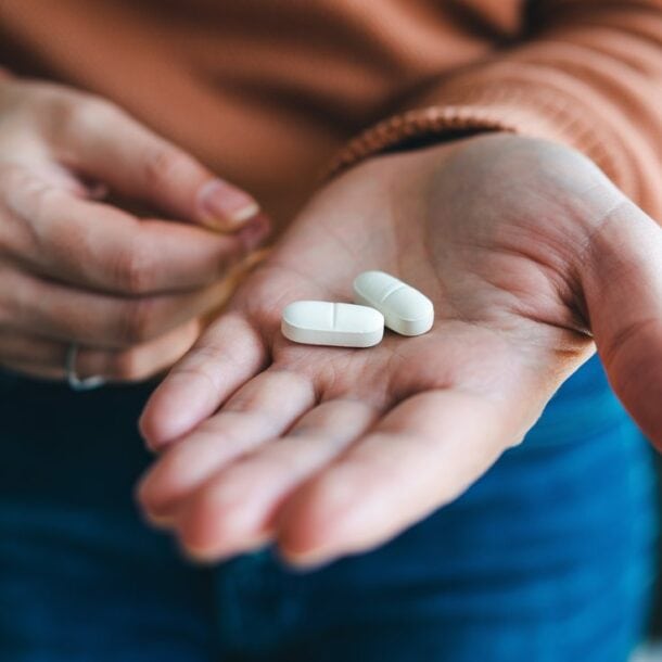 A person holds two large white Foundayo weight loss pills in the palm of their hand, with their other hand reaching toward the pills. The individual is wearing a brown sweater and blue jeans.