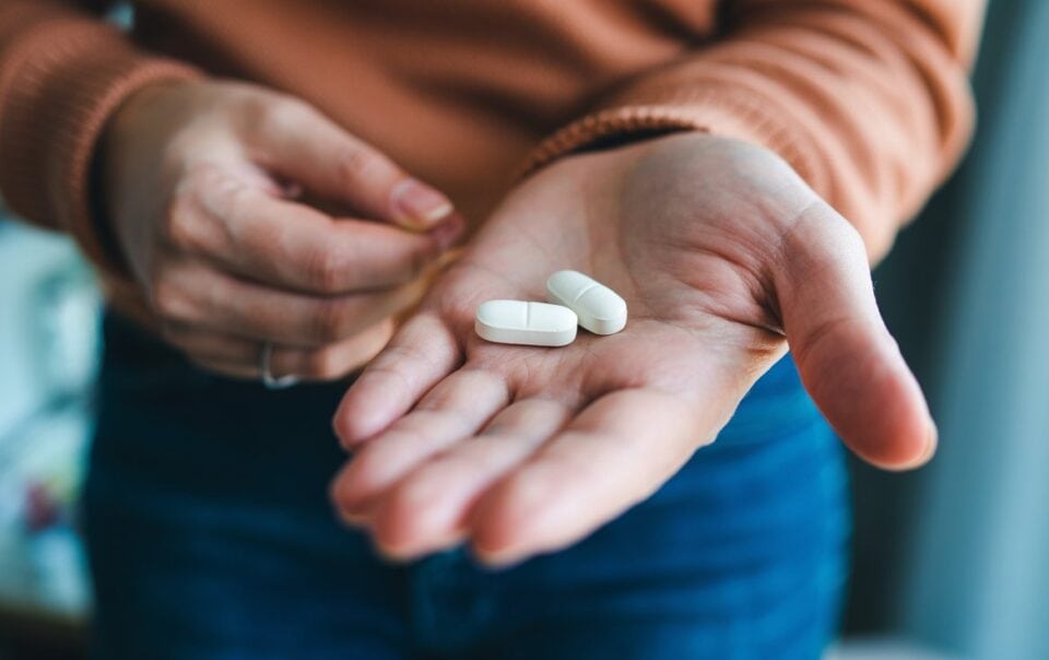 A person holds two large white Foundayo weight loss pills in the palm of their hand, with their other hand reaching toward the pills. The individual is wearing a brown sweater and blue jeans.