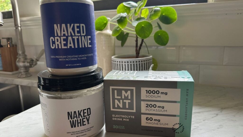 Three supplement containers—Naked Creatine, Naked Whey, and LMNT electrolyte drink mix—sit on a kitchen counter next to a small potted plant, ready to be your Morning Primer Drink for an energy boost. In the background, a sink and window complete the scene.