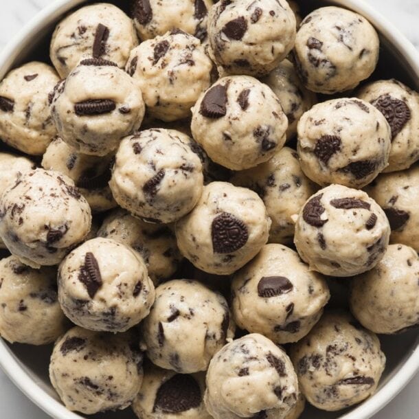 A bowl filled with round cookies and cream protein balls mixed with chunks of chocolate, placed on a white marble surface.