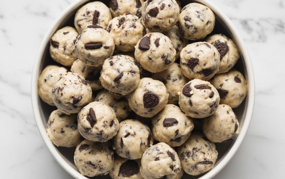 A bowl filled with round cookies and cream protein balls mixed with chunks of chocolate, placed on a white marble surface.