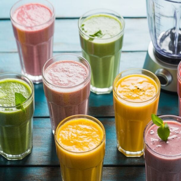 A variety of colorful smoothies in glasses, including green, yellow, pink, and purple, are arranged on a blue wooden table beside a blender. Some are garnished with mint leaves, perfect for anyone following a smoothie guide or seeking a fat burning smoothie.