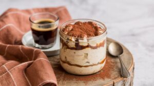 A glass jar of tiramisu overnight oats topped with cocoa powder sits on a round wooden board next to a spoon, a glass of espresso on a saucer, and a brown checkered cloth for the perfect coffee-inspired breakfast.