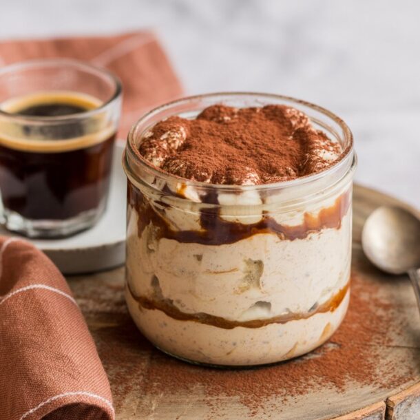 A glass jar of tiramisu overnight oats topped with cocoa powder sits on a round wooden board next to a spoon, a glass of espresso on a saucer, and a brown checkered cloth for the perfect coffee-inspired breakfast.