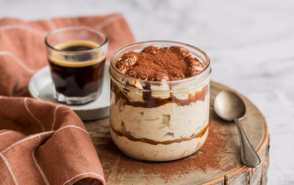 A glass jar of tiramisu overnight oats topped with cocoa powder sits on a round wooden board next to a spoon, a glass of espresso on a saucer, and a brown checkered cloth for the perfect coffee-inspired breakfast.