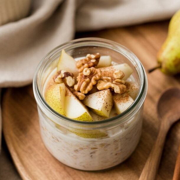 A jar of High Protein Pear Walnut Overnight Oats topped with sliced pears and walnuts sits on a wooden tray. Nearby are a whole pear, a bowl of oats, wooden spoons, and a beige cloth napkin.