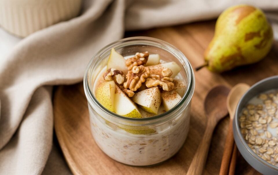 A jar of High Protein Pear Walnut Overnight Oats topped with sliced pears and walnuts sits on a wooden tray. Nearby are a whole pear, a bowl of oats, wooden spoons, and a beige cloth napkin.
