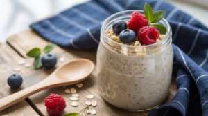 A glass jar filled with overnight oats topped with fresh raspberries, blueberries, and mint leaves sits on a wooden table beside a wooden spoon, scattered oats, and a blue-striped cloth.